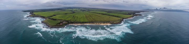 Aerial panorama of a green farmland headland ringed by white surf under an overcast sky