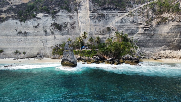 Aerial view of a rock stack and a cluster of palms on a white beach beneath pale limestone cliffs, with turquoise surf