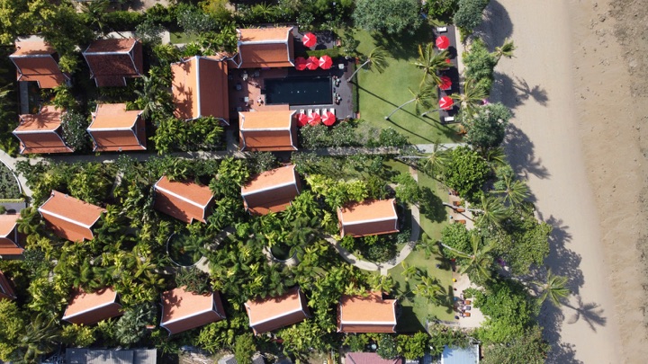 Top-down aerial of terracotta-roofed villas in tropical gardens around a dark pool with red parasols, beside a beach