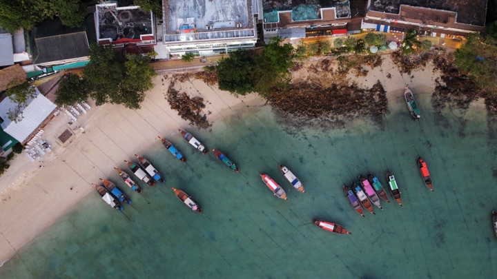Top-down aerial of colourful longtail boats moored in turquoise shallows along a sandy village shoreline