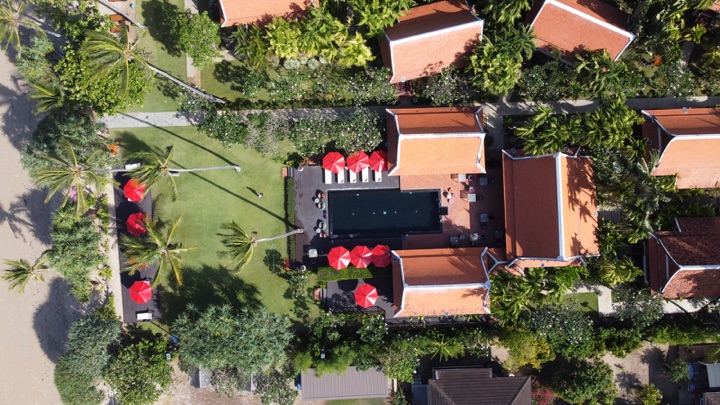Top-down aerial of a dark resort pool ringed by red parasols between terracotta-roofed villas and a palm-dotted lawn