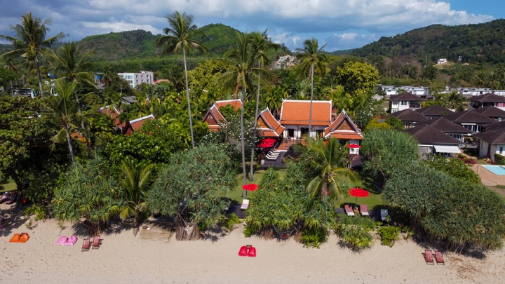 Low aerial view of a Thai-style beachfront resort with orange roofs behind palms and a sandy beach, backed by green hills