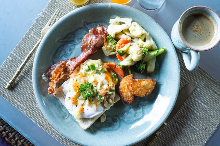 Top-down view of eggs benedict with crispy bacon on a speckled blue plate next to a cup of coffee