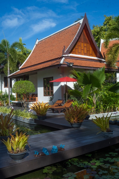 Thai-style villa with an orange tiled roof, a red parasol and loungers on a deck beside a lily pond