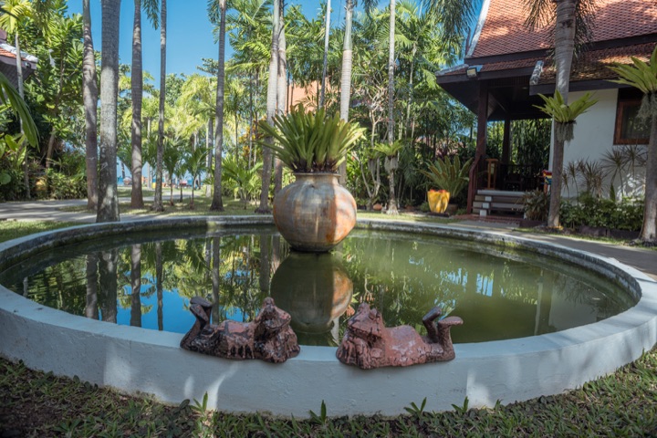 Round garden pond with a large ceramic urn and terracotta figures, reflecting tall palms