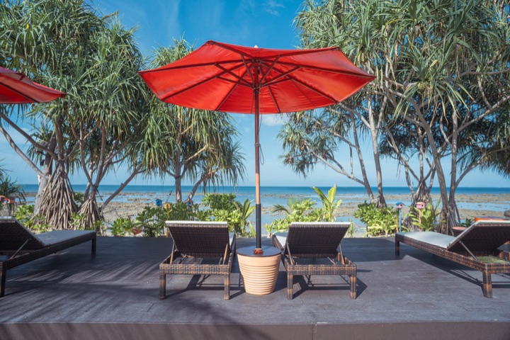 Rattan sun loungers under a red parasol on a deck facing the sea through pandanus trees