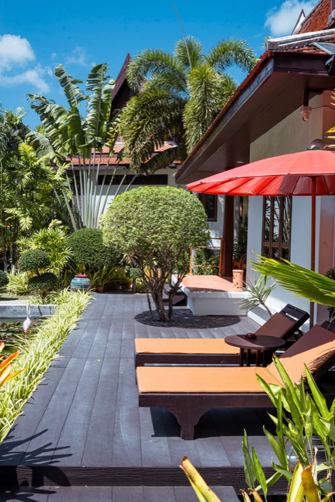 Sun loungers and a red parasol on a dark wooden terrace beside a villa, with clipped trees and palms