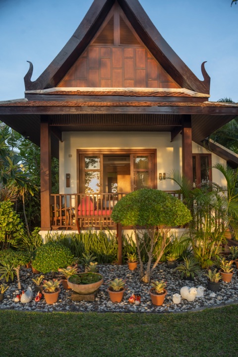 Thai-style villa with a steep carved gable and a veranda in warm low sun, fronted by potted plants
