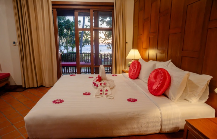 Hotel bed with towel swans and flower petals, with balcony doors opening onto a sea view at dusk