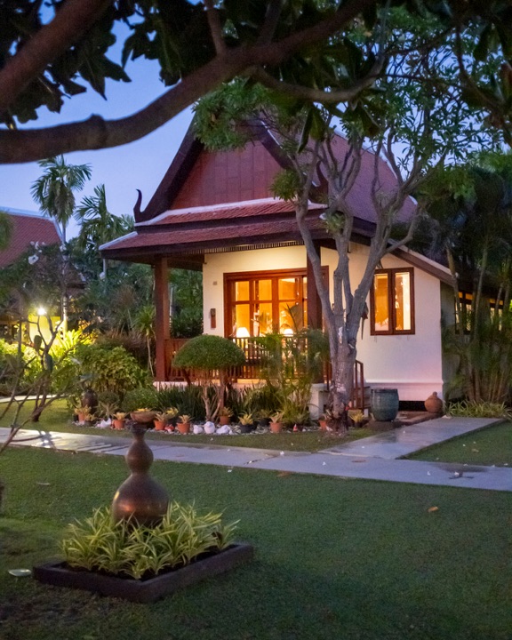 Thai-style villa glowing with warm light at blue hour, framed by frangipani branches and a garden lawn