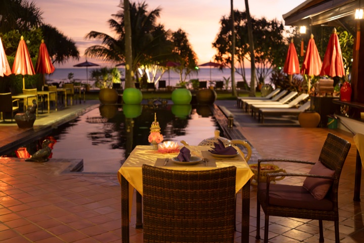 Table set for dinner beside a pool at sunset, with closed parasols and palms against a pink sky