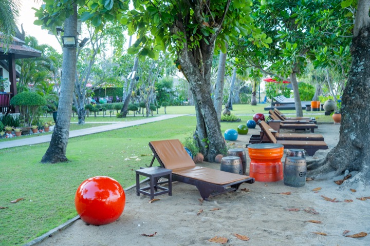 Sun loungers and colourful ceramic stools on sand beneath shady trees beside a resort lawn