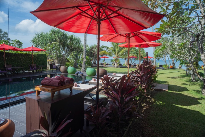 Red umbrellas shading loungers on a lawn beside a pool, with palms and the ocean behind