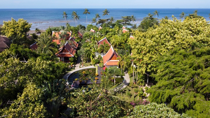 Aerial view over a resort's lotus pond and Thai-style villas among tropical trees, looking out to the sea
