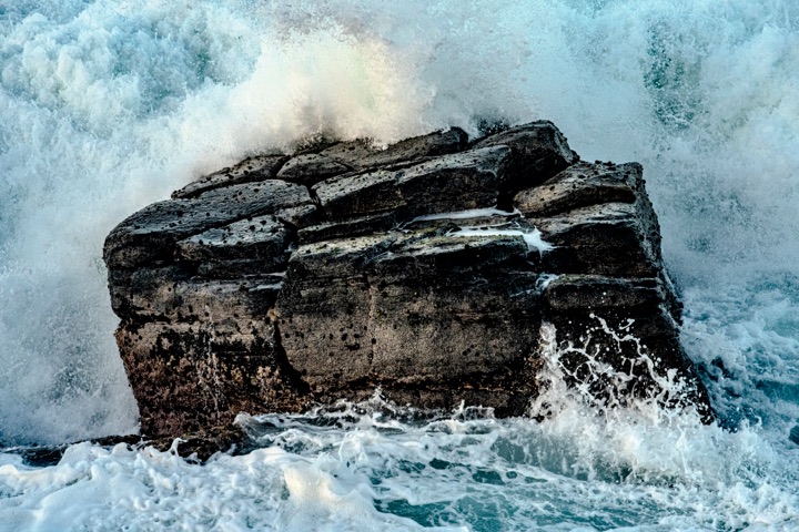 A wave exploding in white spray over a dark volcanic rock