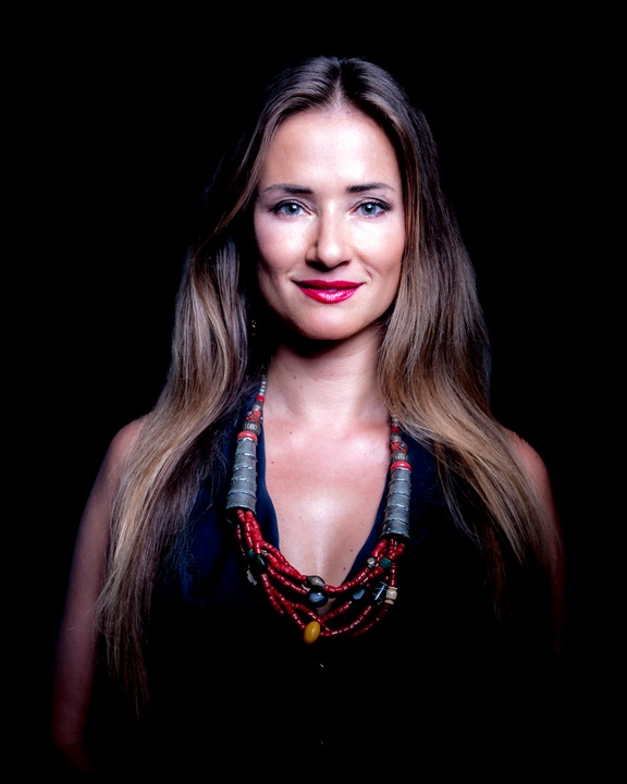 Smiling woman with long hair and a red beaded necklace against a black studio backdrop