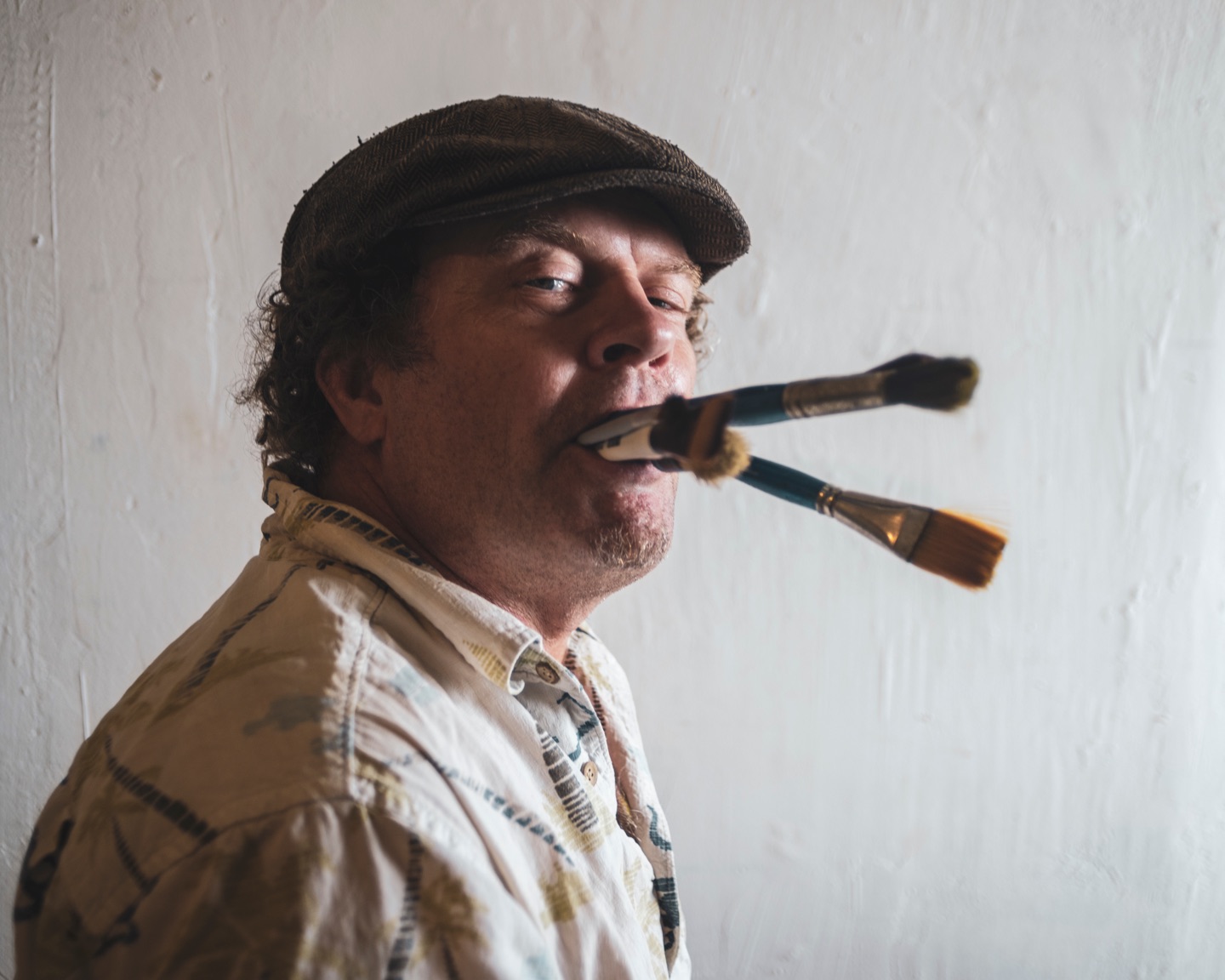 Man in a flat cap holding two paintbrushes in his mouth