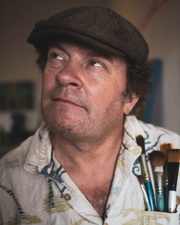 Man in a flat cap and floral shirt looking upward with paintbrushes in his pocket
