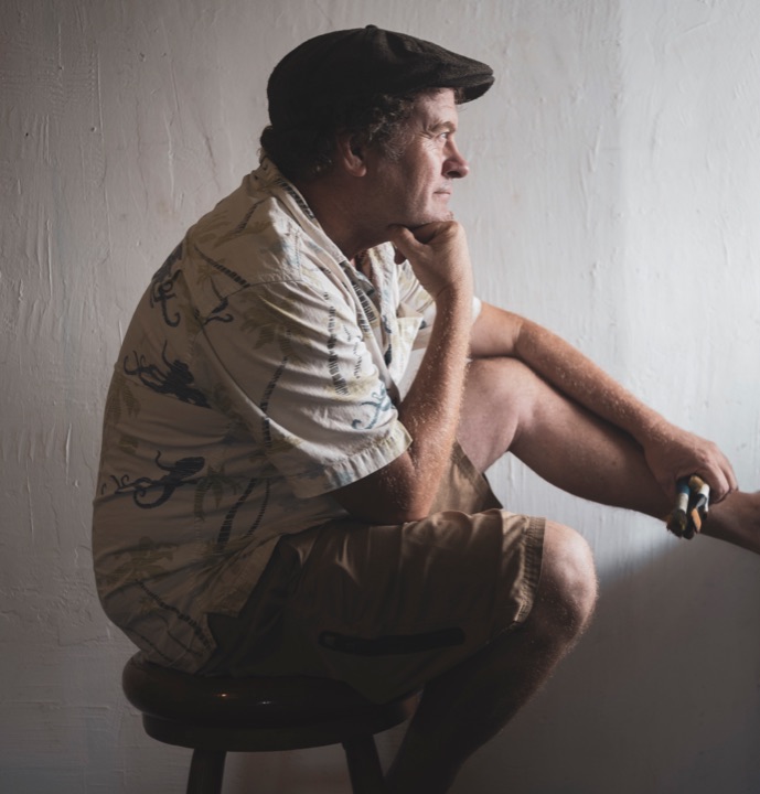 Man in a flat cap sitting thoughtfully on a stool beside a window