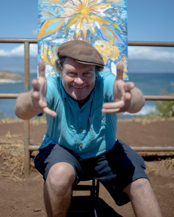 Man in a flat cap framing his face with his hands in front of a colourful painting