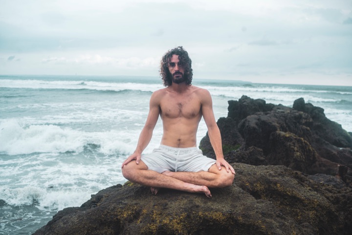 Man meditating cross-legged on rocks beside the ocean
