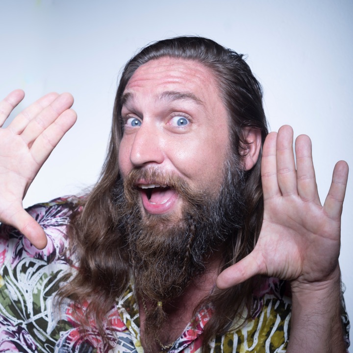 Bearded man in a floral shirt pulling a surprised face with raised hands