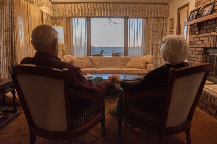 Elderly couple seated side by side in a living room, backlit by a window